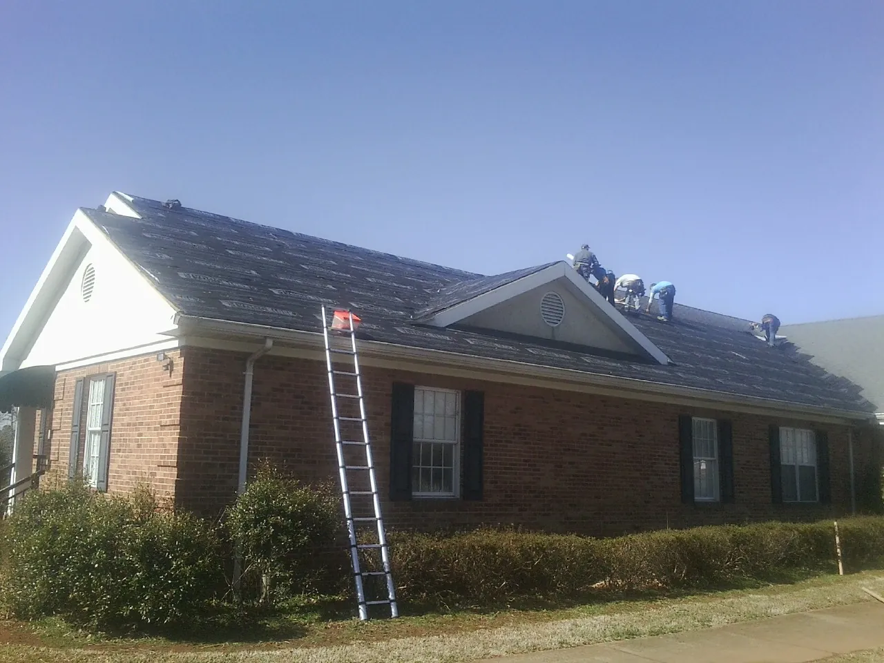 Crew tearing shingles off a brick ranch roof with a ladder set — Emergency Roof Repair work by our crew in Patriot