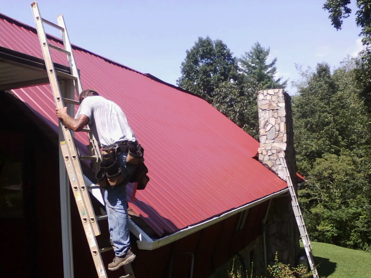 Roofer on a ladder beside a red metal roof and stone chimney — Soffit & Fascia Repair by Roofing Brothers in Patriot