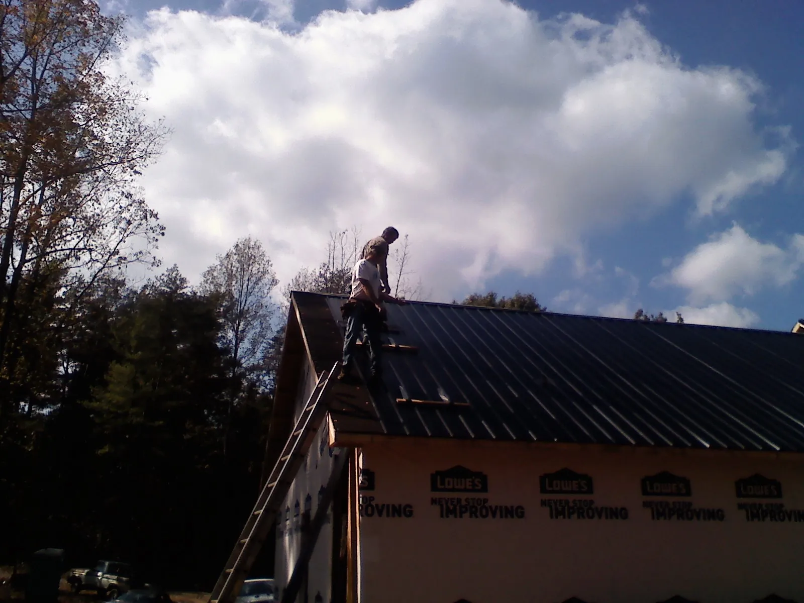Roofer fastening dark metal panels high against a cloudy sky, from a Roof Inspection job in Patriot